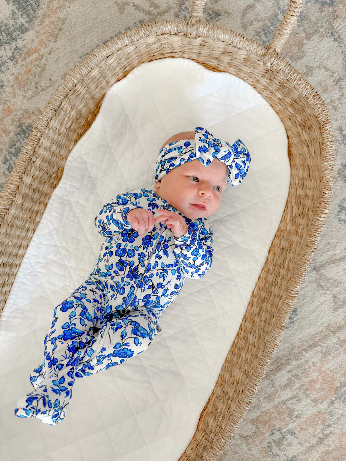 Baby in a blue floral bamboo zippy and headband lying in a woven basket on a stone surface.