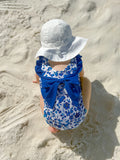 Child in a blue floral swimsuit with a large bow sitting on a sandy beach.