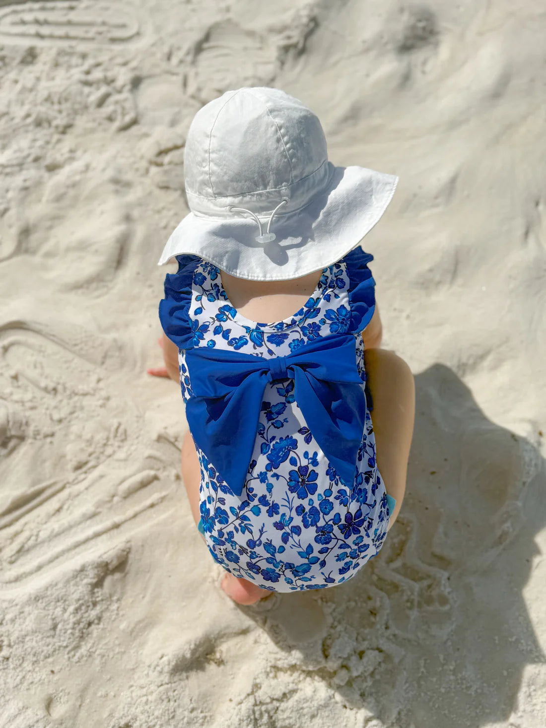 Child in a blue floral swimsuit with a large bow sitting on a sandy beach.