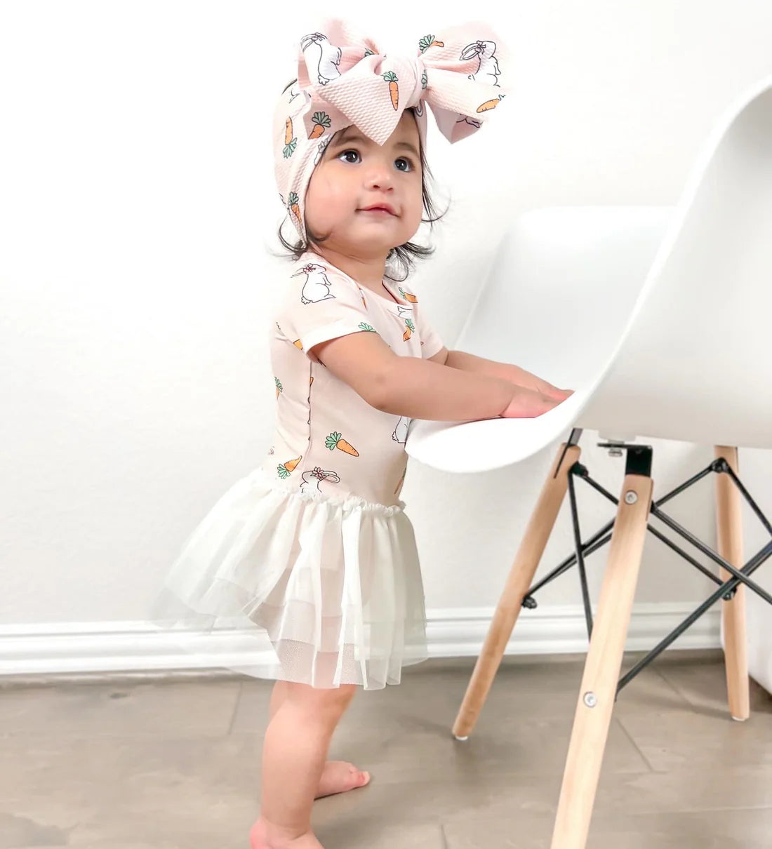 Child wearing a dress with carrot patterns and a large bow headband, standing in a minimalistic room.